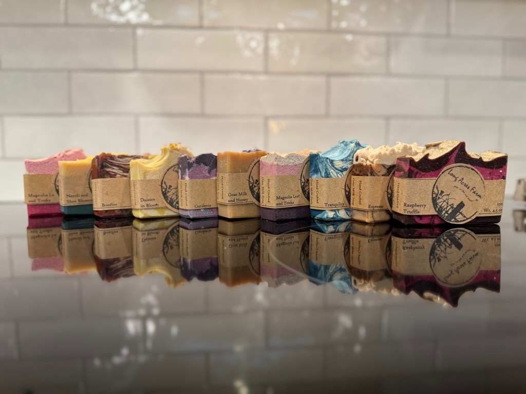 A variety of artisanal soaps displayed in a row, each with unique colors and labels, reflecting different fragrances such as 'Magnolia Leaf and Tonka' and 'Raspberry Truffle'. The soaps are arranged on a shiny surface that reflects their appearance.